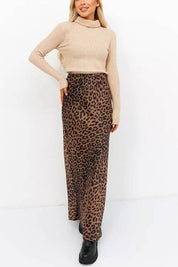 Woman wearing Livia Leopard Satin Skirt with a beige sweater and black boots, showcasing its flowing silhouette and bold print.