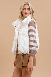 Woman wearing a white Dakota Quilted Puffer Vest with a high zip-up collar, styled for layering over a striped sweater and brown skirt.