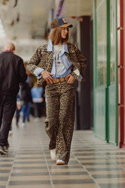 Model showcasing Lynx Denim Pants in bold leopard print paired with a denim shirt and cap, styled in a modern setting.